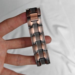 Op de afbeelding: Een koper- en zilverkleurige metalen armband met een geometrisch ontwerp. De armband bestaat uit rechthoekige schakels met een getextureerd oppervlak. De schakels zijn verbonden door kleinere, rechthoekige stukken. De armband wordt tegen een witte achtergrond gehouden.