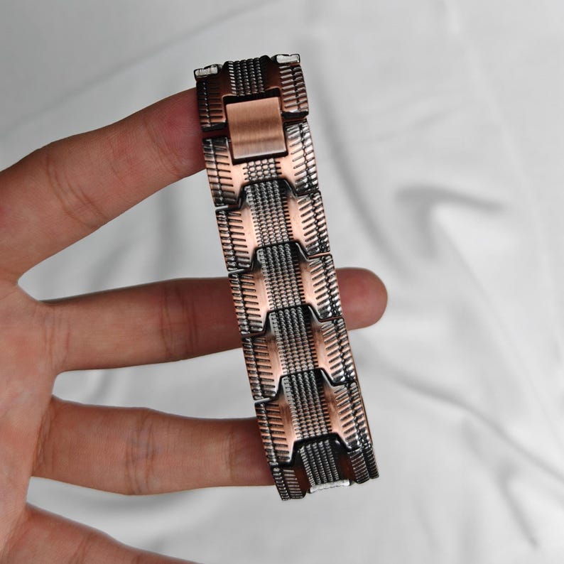 Op de afbeelding: Een koper- en zilverkleurige metalen armband met een geometrisch ontwerp. De armband bestaat uit rechthoekige schakels met een getextureerd oppervlak. De schakels zijn verbonden door kleinere, rechthoekige stukken. De armband wordt tegen een witte achtergrond gehouden.