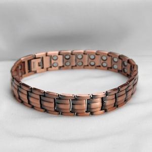 Könnte beinhalten: Ein kupferfarbenes Armband mit rechteckigem Gliederdesign. Das Armband hat einen Magnetverschluss und ist aus Metall gefertigt. Die Glieder sind eine Mischung aus hellen und dunklen Kupfertönen, wodurch ein strukturiertes Aussehen entsteht. Das Armband wird auf einer weißen Oberfläche präsentiert.