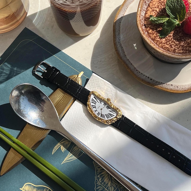 May include: A gold-toned wristwatch with a white face and black Roman numerals, featuring a unique melting design. The watch has a black leather band. The watch is displayed on a white napkin.