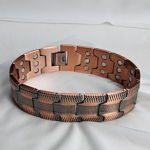 Op de afbeelding: Een koperkleurige armband met een magnetische sluiting. De armband bestaat uit rechthoekige schakels, sommige effen en andere met patronen. De schakels zijn verbonden door scharnieren, wat flexibiliteit biedt. De armband is ontworpen om om de pols te worden gedragen.
