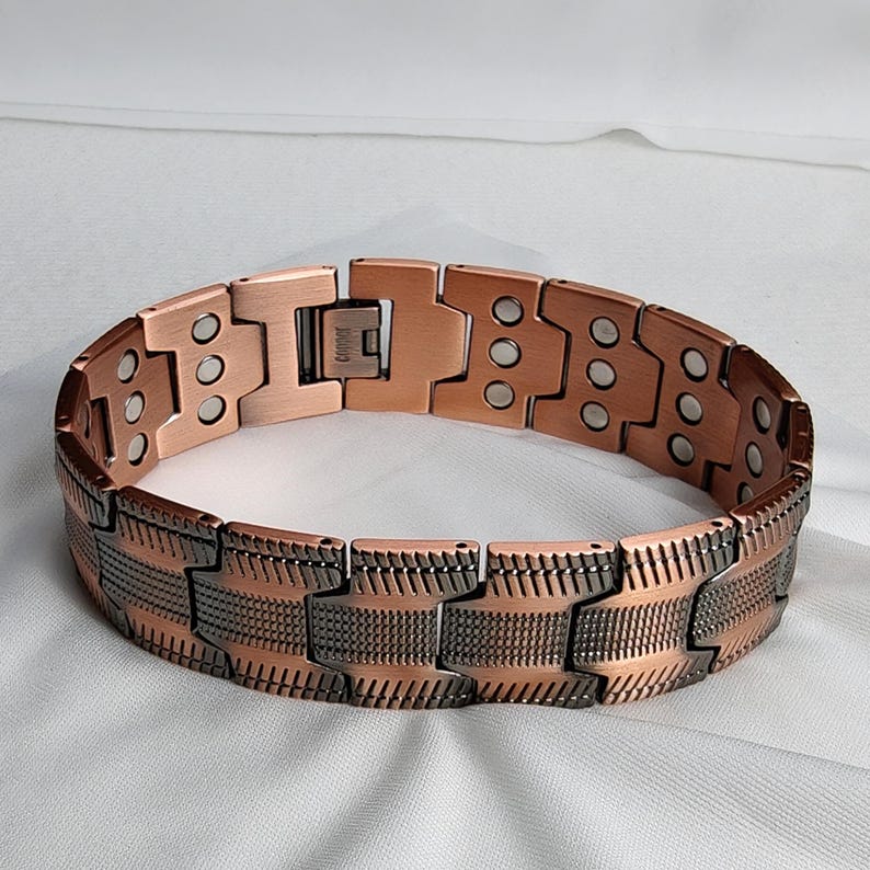 Op de afbeelding: Een koperkleurige armband met een magnetische sluiting. De armband bestaat uit rechthoekige schakels, sommige effen en andere met patronen. De schakels zijn verbonden door scharnieren, wat flexibiliteit biedt. De armband is ontworpen om om de pols te worden gedragen.