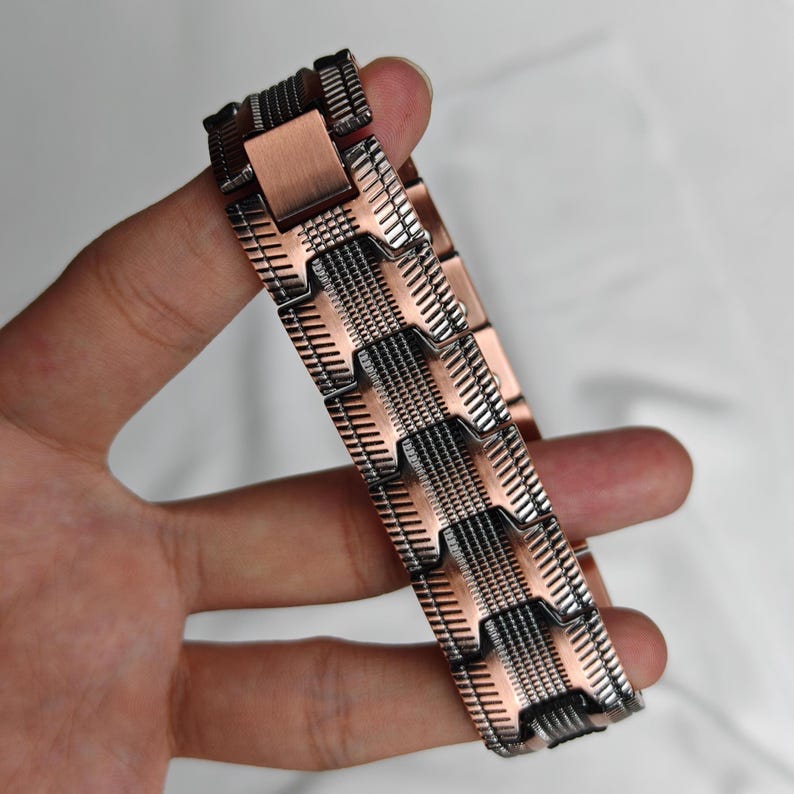 Op de afbeelding: Een koperkleurige metalen armband met een geometrisch ontwerp. De armband wordt in een hand gehouden en heeft een reeks rechthoekige en patroonvormige schakels. Het metaal heeft een mix van koper- en donkerdere tinten.