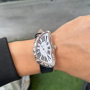 May include: A unique wristwatch with a white face and black Roman numerals. The watch has a silver-colored, textured bezel and a black leather band. The watch face has a melting design, giving it a surreal appearance.