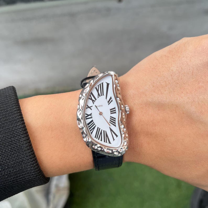 May include: A unique wristwatch with a white face and black Roman numerals. The watch has a silver-colored, textured bezel and a black leather band. The watch face has a melting design, giving it a surreal appearance.