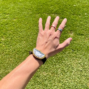 May include: A gold-toned watch with a white face and black Roman numerals. The watch has a unique, asymmetrical shape and a black leather band. Several rings are visible on the fingers.