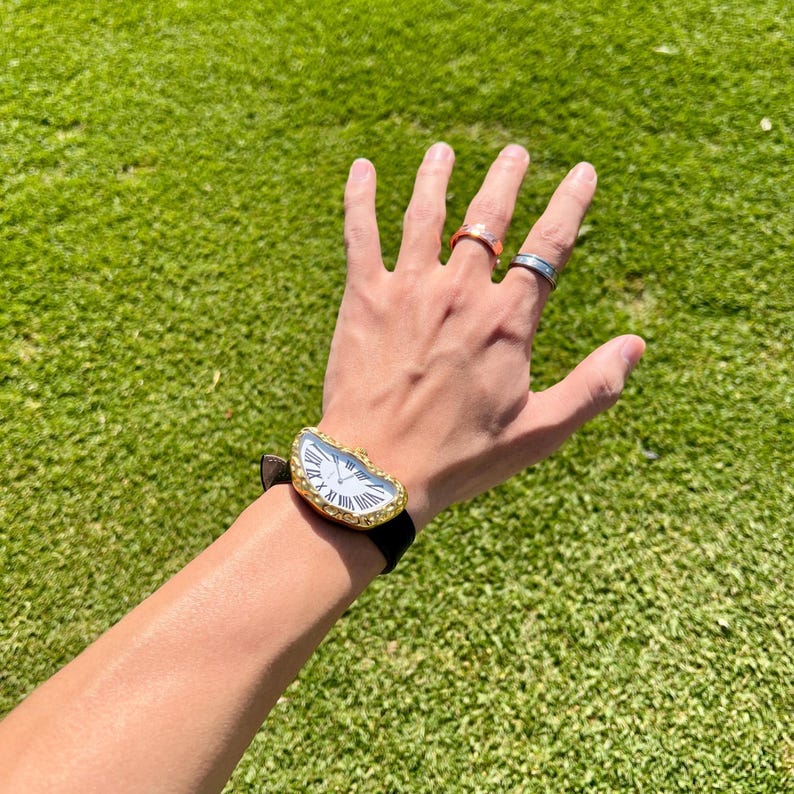 May include: A gold-toned watch with a white face and black Roman numerals. The watch has a unique, asymmetrical shape and a black leather band. Several rings are visible on the fingers.