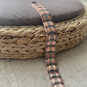 Op de afbeelding: Een koperkleurige armband met een geometrisch ontwerp. De armband heeft rechthoekige schakels met een gestructureerd, gestreept patroon in een donkerdere metallic kleur. De armband wordt gepresenteerd op een geweven, cirkelvormig oppervlak.