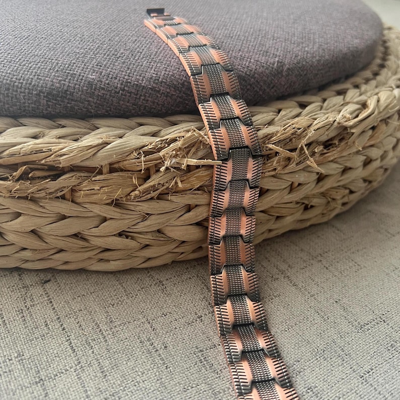 Op de afbeelding: Een koperkleurige armband met een geometrisch ontwerp. De armband heeft rechthoekige schakels met een gestructureerd, gestreept patroon in een donkerdere metallic kleur. De armband wordt gepresenteerd op een geweven, cirkelvormig oppervlak.