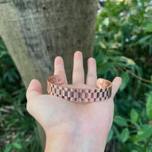 May include: Copper-colored cuff bracelet with a rectangular link design. The bracelet is open-ended and appears to be made of metal. The bracelet is held in a hand, with a blurred background of green foliage and a tree trunk.