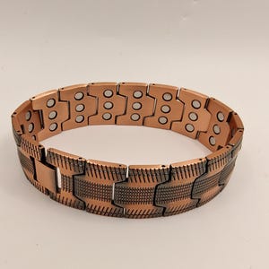 Op de afbeelding: Een koperkleurige magnetische armband met een gesegmenteerd ontwerp. De armband heeft rechthoekige schakels met cirkelvormige inkepingen en een gestructureerd patroon op het onderste gedeelte. Het metaal heeft een warme, roodbruine tint.