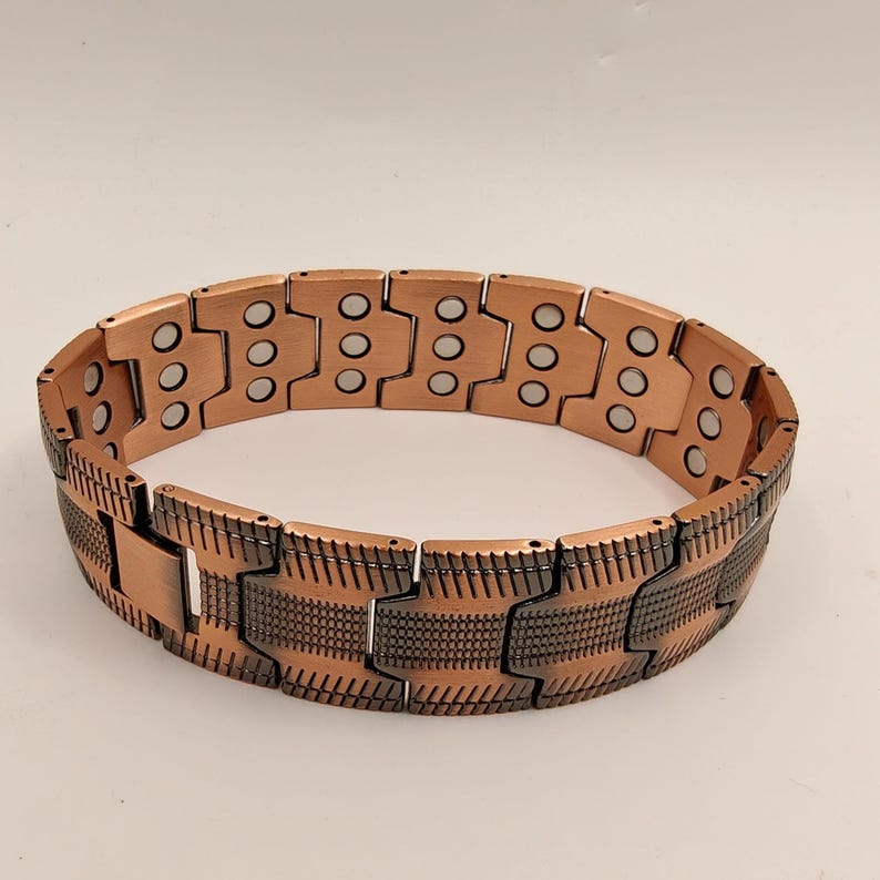 Op de afbeelding: Een koperkleurige magnetische armband met een gesegmenteerd ontwerp. De armband heeft rechthoekige schakels met cirkelvormige inkepingen en een gestructureerd patroon op het onderste gedeelte. Het metaal heeft een warme, roodbruine tint.