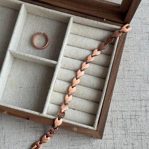 May include: A copper-colored bracelet with a leaf-like design and a matching ring are displayed inside a wooden jewelry box with a glass lid. The box has a beige fabric lining.