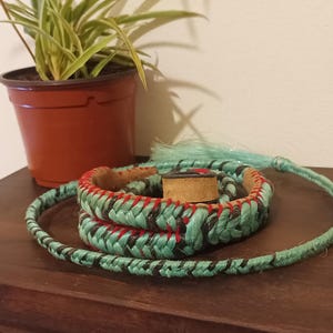Pode incluir: Um cinto trançado turquesa e preto com costuras vermelhas e um detalhe em couro, enrolado sobre uma superfície de madeira. Uma pequena planta em vaso está ao fundo.