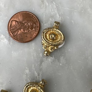 May include: Five gold-toned metal pendant charms with ornate designs. Each charm features a circular center with a raised, textured surface and a decorative border. The charms have a loop at the top for attaching to a necklace or bracelet.