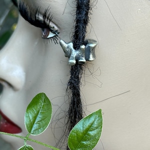 May include: A silver-toned dreadlock bead in the shape of a small dinosaur is featured in this close-up shot. The bead is attached to a black dreadlock. The background includes a mannequin's face with long eyelashes and red lipstick, and green leaves.