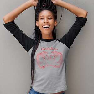 May include: A gray raglan shirt with black sleeves featuring a red graphic and the text "fiercelocks.com". The shirt is paired with blue jeans. The model is smiling with their arms raised.