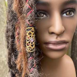 May include: A gold hair tie with a decorative design and a pink gem in the center. The hair tie is worn in a person's hair.