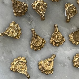 May include: A collection of 10 gold-toned metal pendants with intricate designs. Each pendant features a circular pattern with a raised center and a beaded border. The pendants have a teardrop shape and a loop at the top for attaching to a chain or cord.