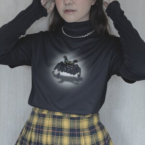 Może przedstawiać: Czarny, długorękawny t-shirt z nadrukiem pająka z przodu. Koszulka jest połączona z ż&oacute;łto-czarną sp&oacute;dnicą w kratę.