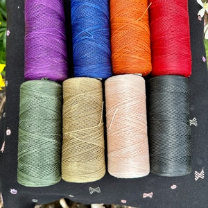 May include: Eight spools of waxed thread in various colors, including purple, blue, orange, red, green, beige, pink, and black. The spools are arranged in two rows of four on a black fabric background.