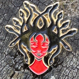 May include: A red and black enamel pin depicting a woman with snakes for hair. The woman has closed eyes and a red face. The snakes are black with gold accents.