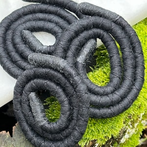 May include: A close-up of a black coiled rope made of fabric. The rope is coiled in a spiral shape and is sitting on a bed of green moss.