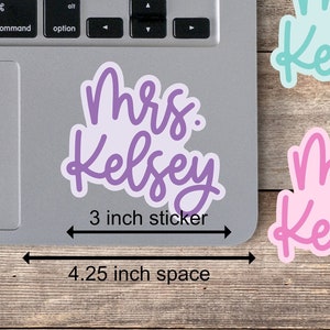 May include: Three 3-inch stickers with the text "Mrs. Kelsey" in different colors: pink, purple, and blue. The stickers are arranged on a wooden surface with a laptop in the background. The text "4.25 inch space" is written below the stickers.