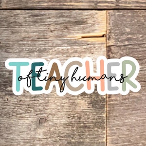 May include: A colorful sticker with the word "TEACHER" in large letters and the phrase "of tiny humans" written in cursive below. The sticker is on a wood background.