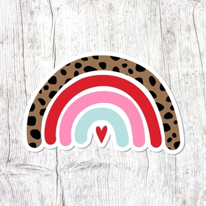 May include: A rainbow sticker with a leopard print pattern and a red heart in the center. The rainbow is made of red, pink, light blue, and white.