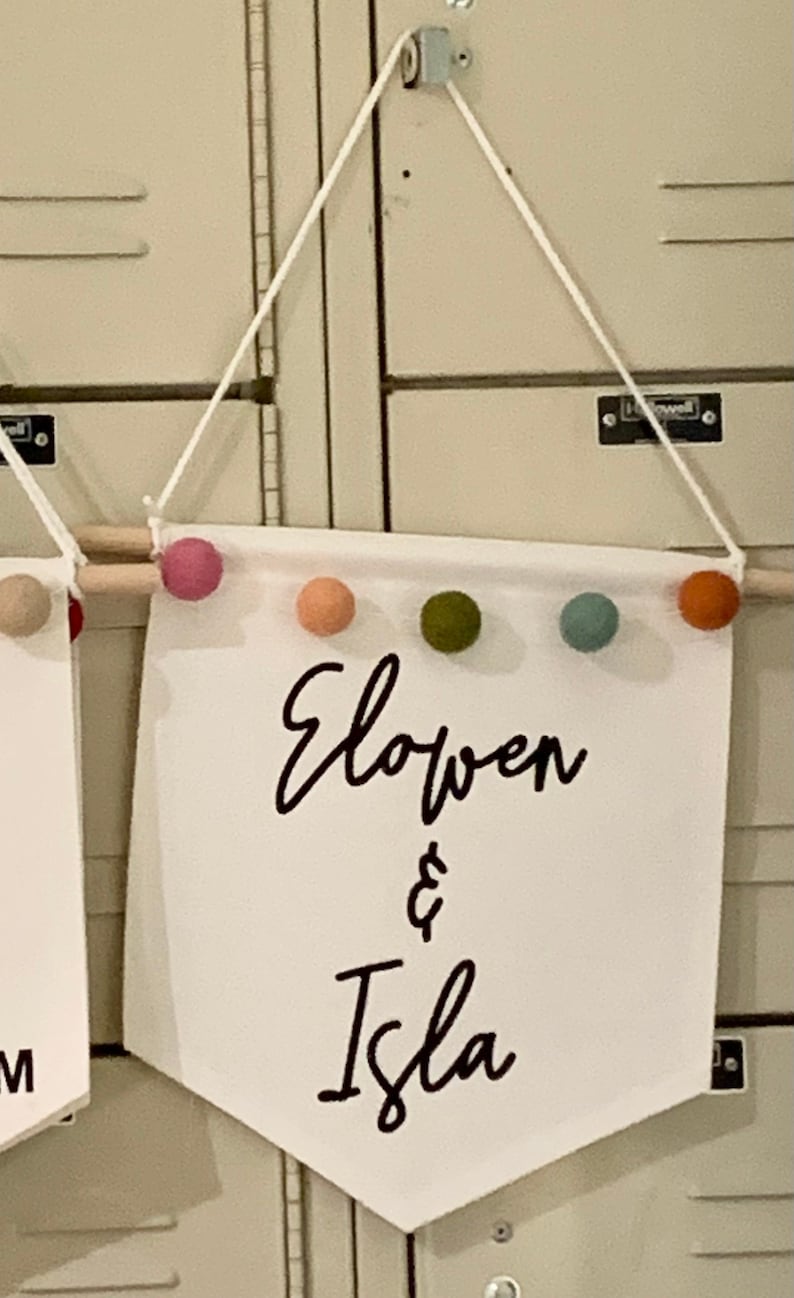 Peut inclure: Banni&egrave;re en tissu blanc avec les noms "Elowen & Isla" &eacute;crits en cursive noire. La banni&egrave;re est d&eacute;cor&eacute;e d'une baguette en bois et de boules de feutre color&eacute;es.