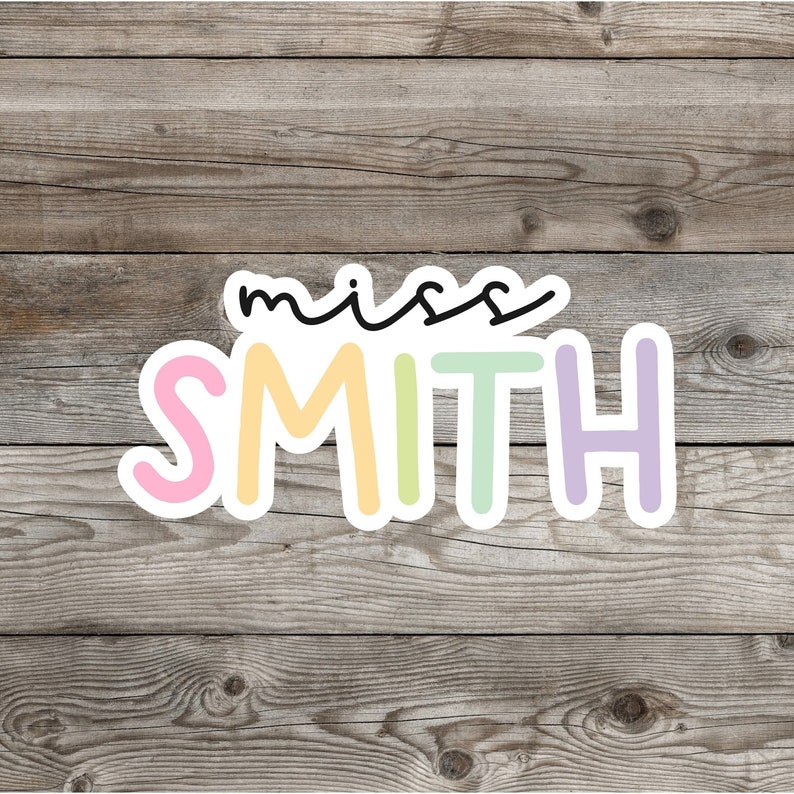 Pu&ograve; includere: Un adesivo con il testo "miss SMITH" in un carattere colorato. Le lettere sono contornate di bianco e hanno uno schema di colori pastello. Lo sfondo &egrave; una texture in legno.