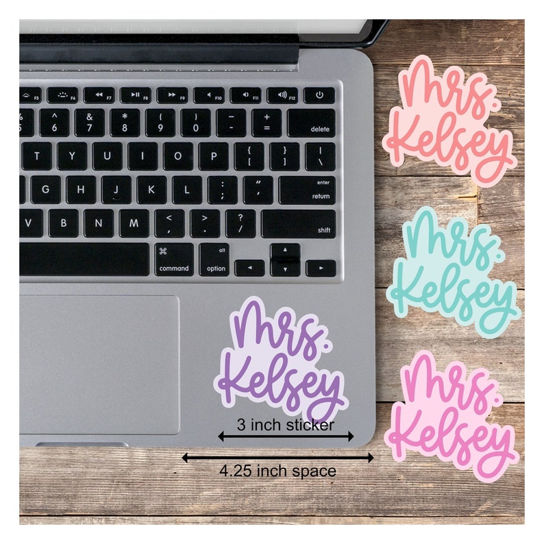 May include: Three 3-inch stickers with the text "Mrs. Kelsey" in different colors: pink, purple, and blue. The stickers are arranged on a wooden surface with a laptop in the background. The text "4.25 inch space" is written below the stickers.