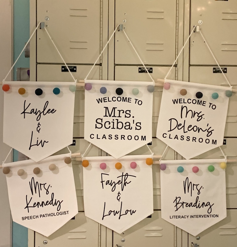 Peut inclure: Cinq banni&egrave;res blanches en forme de fanion avec du texte noir et des pompons color&eacute;s suspendus &agrave; un casier. Les banni&egrave;res indiquent : "Kaylee & Liv", "Bienvenue dans la classe de Mme Sciba", "Bienvenue dans la classe de Mme Deleon", "Mme Kennedy Orthophoniste", et "Fayeth & LouLou".