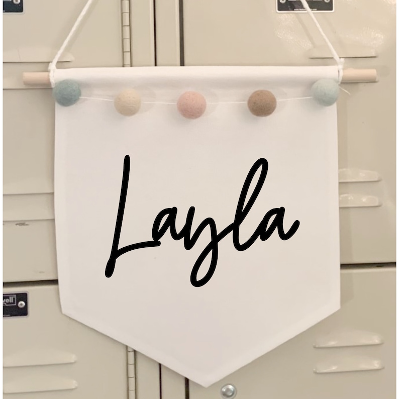 Peut inclure: Banni&egrave;re en tissu blanc avec un nom en &eacute;criture cursive noire "Layla" et une cha&icirc;ne de pompons de couleur pastel en haut.