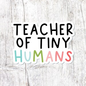 May include: White sticker with the text "Teacher of Tiny Humans" in black. The word "Humans" is in rainbow colors.