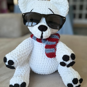 May include: A white crocheted teddy bear wearing black sunglasses and a striped scarf in red, gray, and blue. The bear has black paws and a black nose. The bear is sitting on a neutral-colored surface.