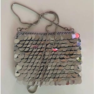 Op de afbeelding: Een zilveren pailletten handtas met een zilveren kettingriem. De tas is rechthoekig met een gehaakte grijze bovenkant. De pailletten zijn rond en overlappen elkaar, waardoor een glinsterend effect ontstaat. De ketting heeft een dun, slangachtig ontwerp.