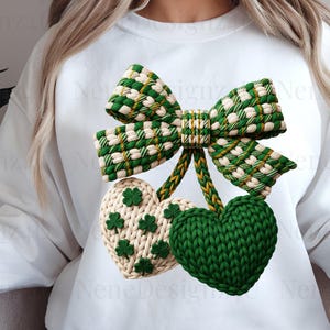 May include: A white sweatshirt featuring a green and white bow and two heart-shaped ornaments. One heart is green, the other is cream-colored with green shamrocks. The design is likely for St. Patrick's Day.