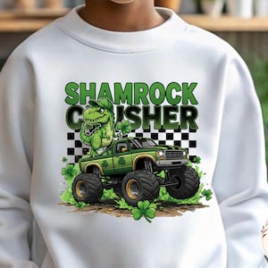 May include: White sweatshirt with a graphic featuring a green monster truck and a dinosaur wearing a hat, with the text "Shamrock Crusher" in green. The design includes shamrocks and a checkered background.