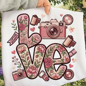 Può includere: Borsa tote bianca con la parola "LOVE" in un design floreale e un'illustrazione di fotocamera rosa. Le lettere sono riempite di fiori rosa e foglie verdi. La borsa include anche rulli di pellicola, cuori e il testo "CAPTURE MOMENTS".