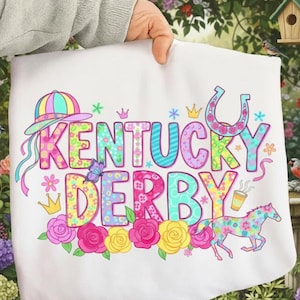 May include: A white tote bag featuring the words "Kentucky Derby" in colorful, floral-patterned letters. The design includes a hat, horseshoe, horse, and roses. The bag is held by a person.