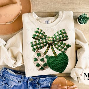 May include: Cream-colored sweatshirt featuring a green and white plaid bow and two heart-shaped designs. One heart is green, the other is cream with green shamrocks. The sweatshirt is paired with blue jeans and a hat.