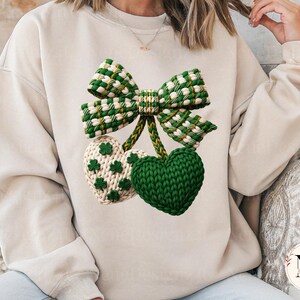 May include: A cream-colored sweatshirt featuring a green and white bow and two heart-shaped designs. One heart is solid green, and the other is cream with green shamrock accents. The bow and hearts appear to be knitted or crocheted.