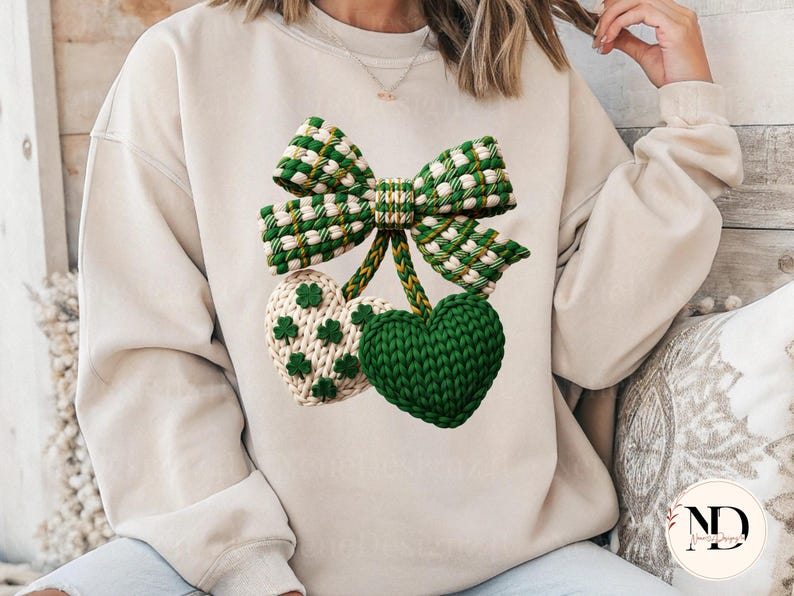 May include: A cream-colored sweatshirt featuring a green and white bow and two heart-shaped designs. One heart is solid green, and the other is cream with green shamrock accents. The bow and hearts appear to be knitted or crocheted.