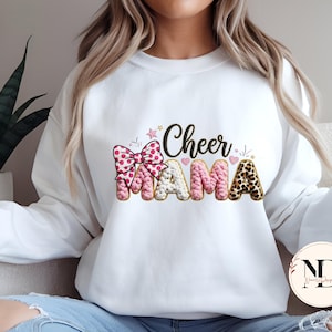 May include: White sweatshirt featuring the text "Cheer Mama". The word "Cheer" is in black script, and "Mama" is in pink, white, and leopard print. The design includes a pink bow and star accents.