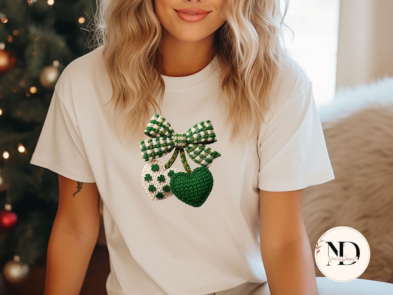 May include: A white t-shirt featuring a green and white gingham bow and two knitted heart-shaped ornaments. One ornament is green, and the other is white with green shamrock patterns. The design is centered on the shirt.