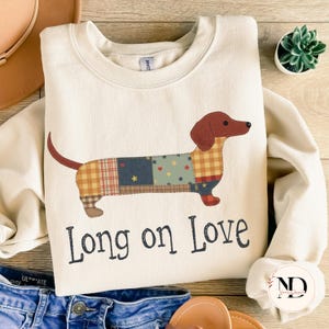 Puede incluir: Sudadera crema con un diseño de perro salchicha de retazos con detalles en marrón, rojo y azul. El texto "Long on Love" está impreso debajo. La sudadera se combina con unos vaqueros azules.