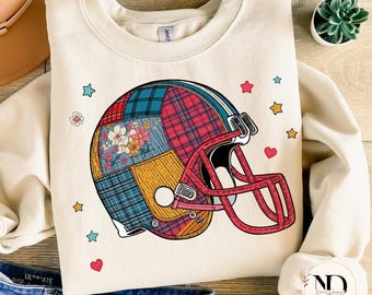 May include: Cream-colored sweatshirt featuring a football helmet design. The helmet is a patchwork of floral, plaid, and solid fabrics in red, blue, and yellow. The helmet has a red face mask. Scattered around the helmet are stars, hearts, and flowers.