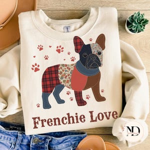 Puede incluir: Sudadera color crema con un diseño de bulldog francés en patchwork. El perro presenta secciones de tela a cuadros, floral y lisa. El texto "Frenchie Love" está impreso en una fuente marrón rústica. Huellas de patas rojas salpican el diseño.